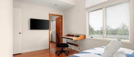 Ein Schlafzimmer mit Bett, Schreibtisch, Stuhl, Fernseher und einem Fenster mit Jalousien.