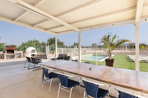 En udendørs terrasse med et træbord og stole, grill og udsigt til et poolområde og grønne omgivelser.