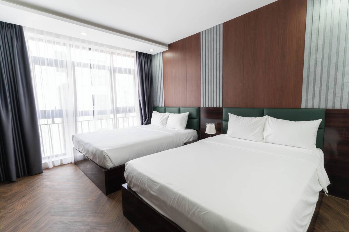 Une chambre d’hôtel avec deux lits simples, un plancher en bois, une grande fenêtre avec des rideaux et une tête de lit en bois.