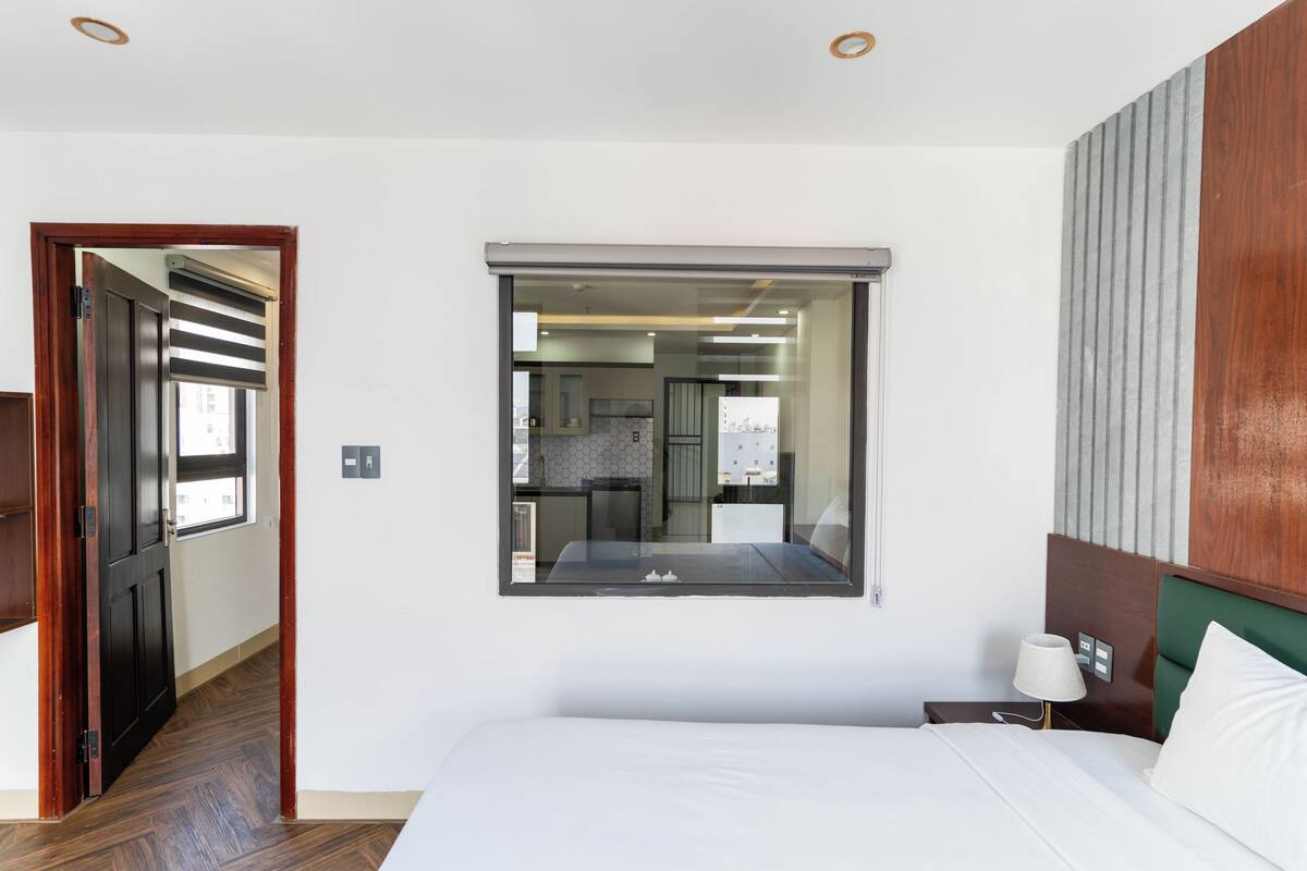 Une chambre d’hôtel avec un grand lit, une table de chevet avec une lampe, un miroir au mur et une vue sur la cuisine qui se reflète dans le miroir.