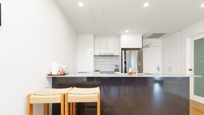Eine moderne Küche mit weißen Schränken, einem Edelstahlkühlschrank und einer dunklen Kücheninsel. Zwei Holzhocker und ein kleiner Tisch sind ebenfalls vorhanden.