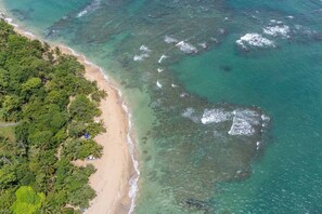 Une vue aérienne d’une plage aux eaux turquoise et limpides, bordée d’un rivage sablonneux et d’une végétation luxuriante.