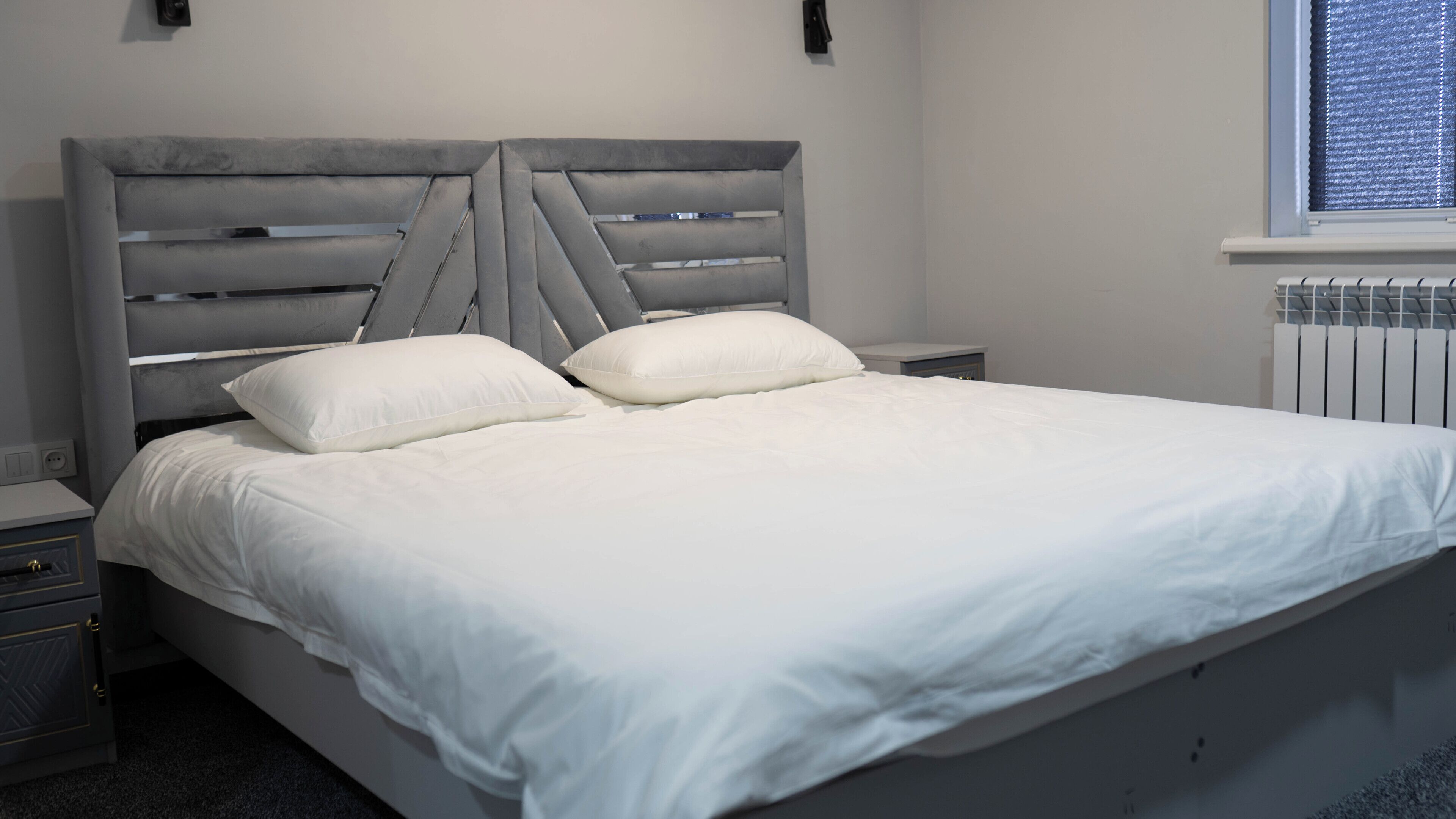A bedroom with a bed, two bedside tables, a window with blinds, and a radiator.