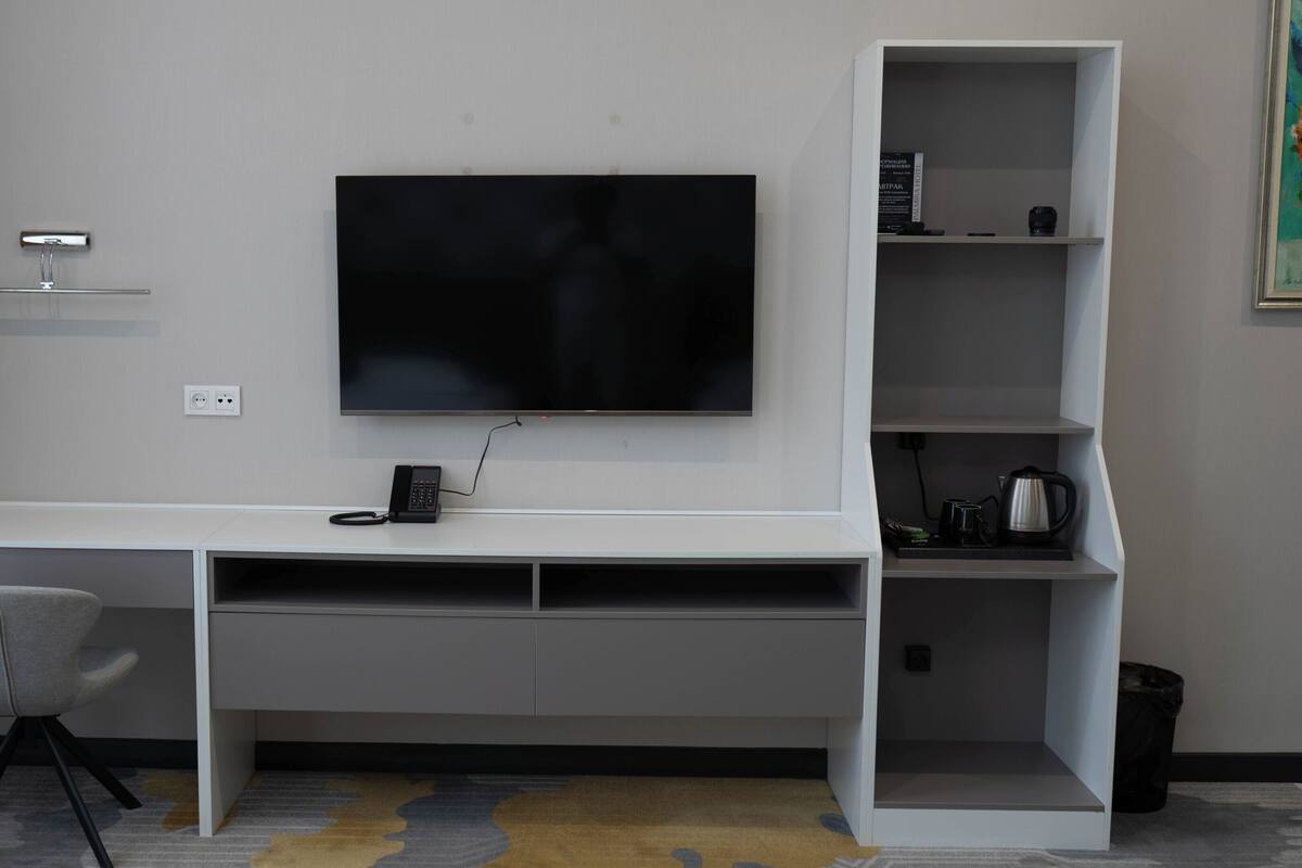 A modern hotel room with a white desk, a mounted flat-screen TV, a chair, and a coffee machine.