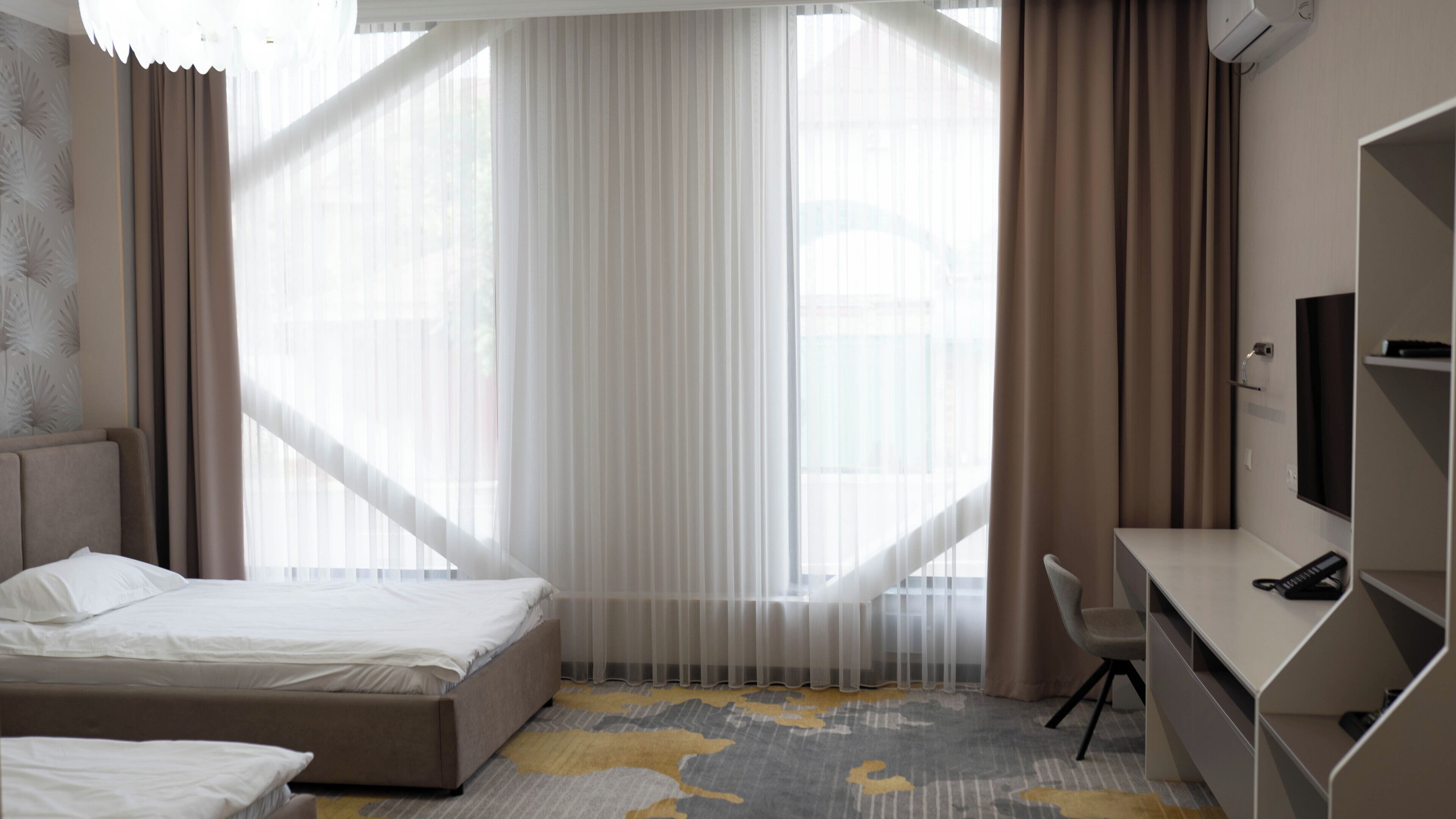 Une chambre d’hôtel avec un lit, un bureau, une chaise et une grande fenêtre avec des rideaux légers.