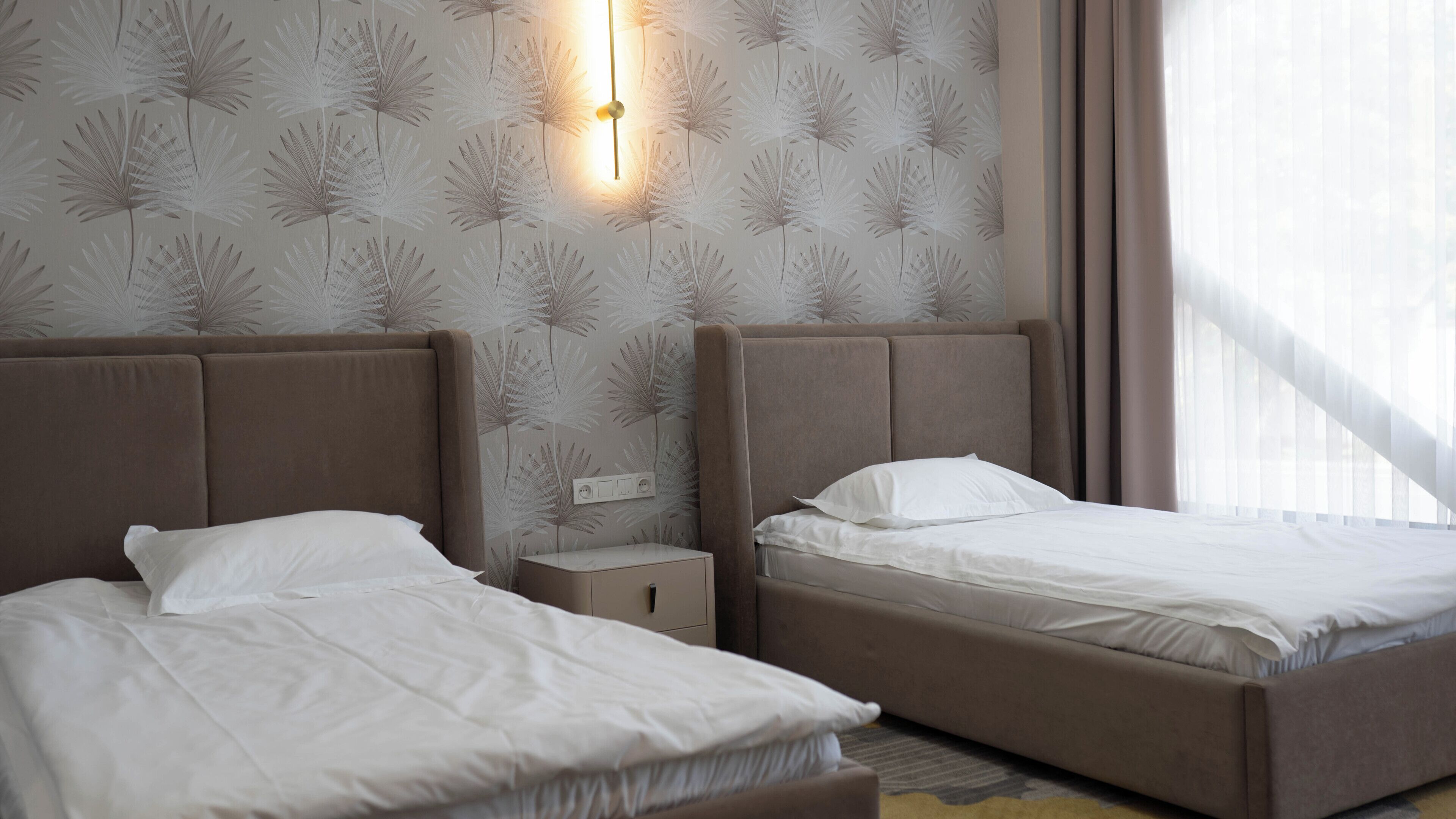 A hotel room with two beds, a nightstand, a wall-mounted light, and a window with curtains.
