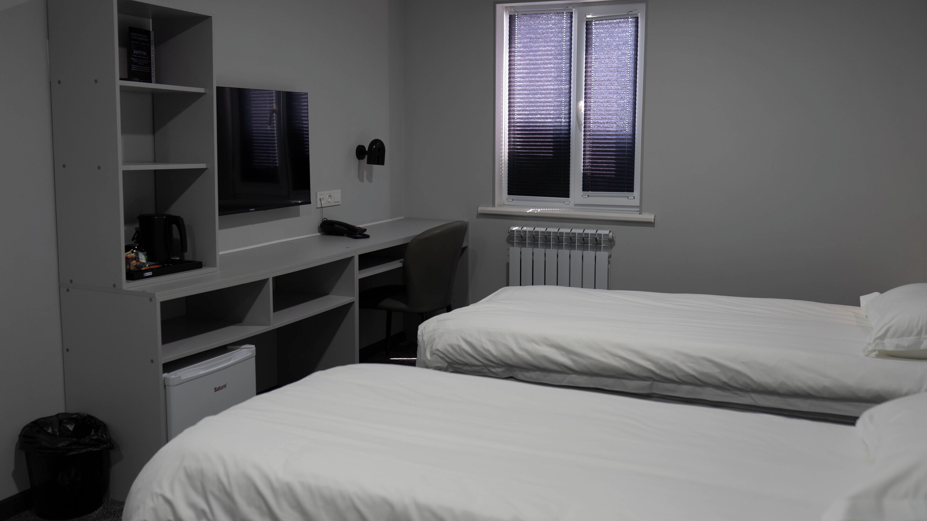 Un dormitorio con dos camas, un escritorio con computadora, un radiador y una ventana con persianas.