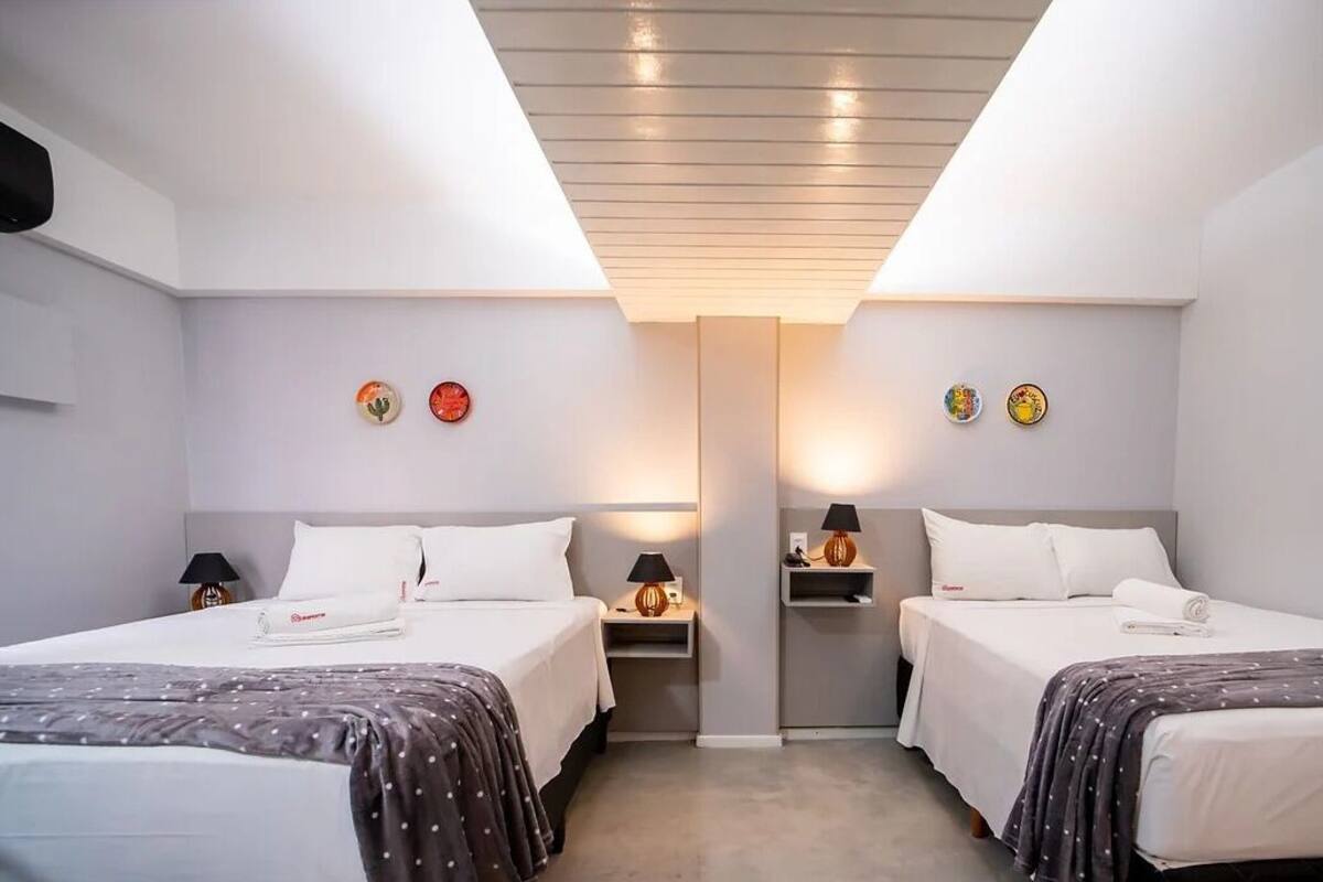 Une chambre d’hôtel avec deux lits, chacun étant équipé d’un oreiller et d’une couverture. On y trouve des tables de chevet avec des lampes et une petite étagère. La chambre présente un design moderne, avec un plafond et des murs de couleur claire.