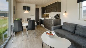 Una cucina moderna con mobili scuri, una zona pranzo con tavolo bianco e sedie nere, un divano grigio e una finestra con vista su una zona verde.