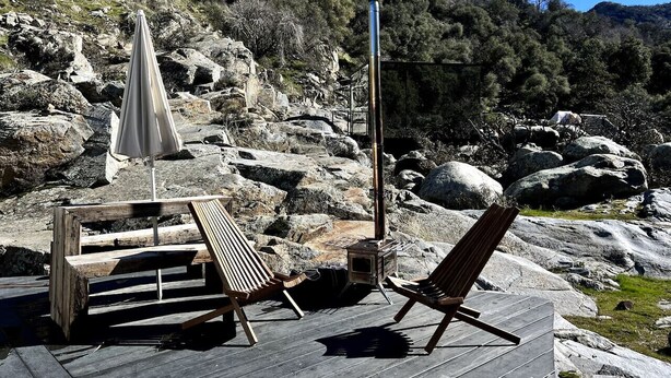 A wooden deck with two lounge chairs and a table, a stone fireplace, and a rocky hillside in the background.