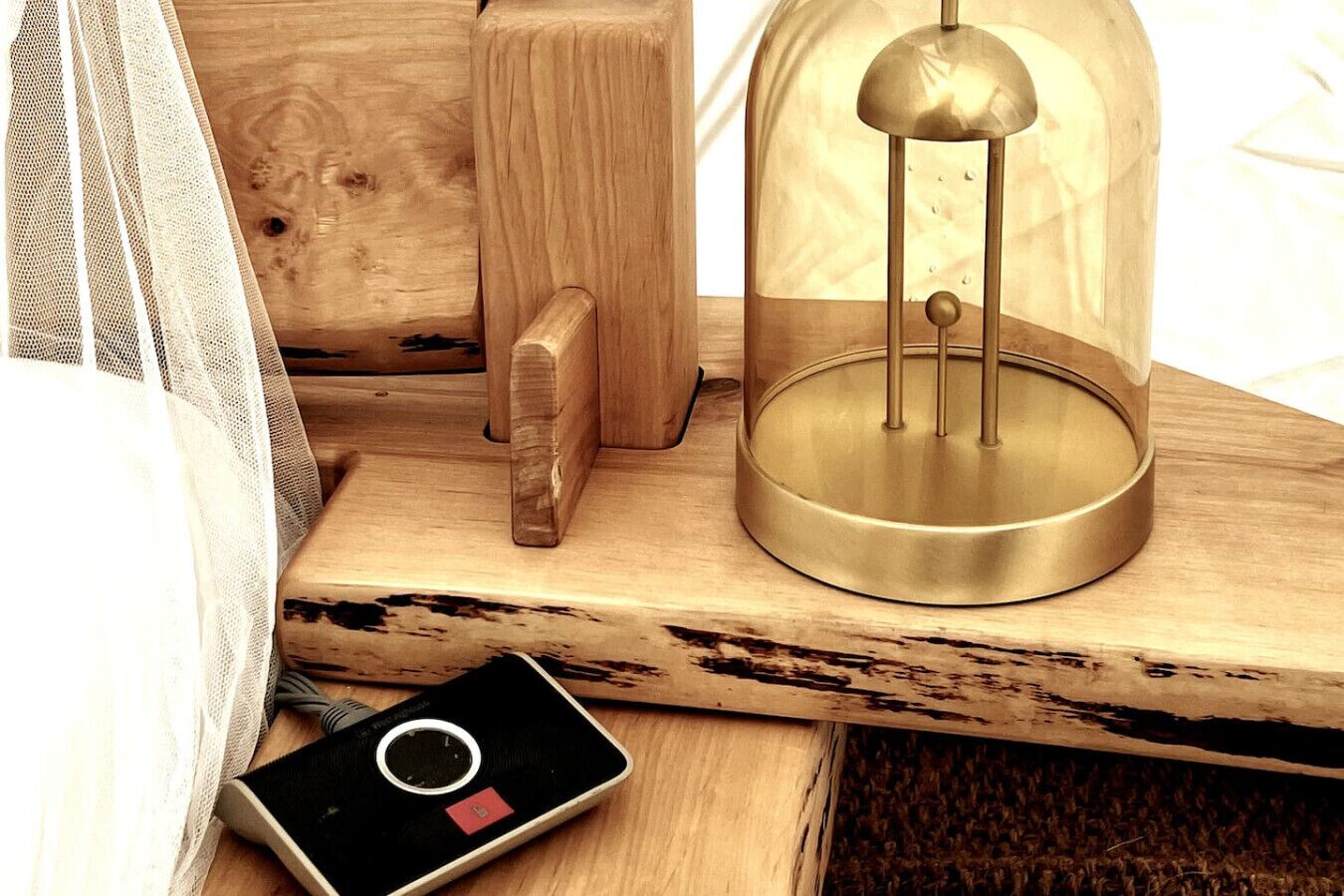 A wooden bedside table with a brass lamp, a small black device, and a white mesh bag.