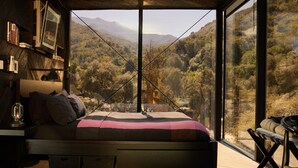 A bedroom with a bed, a nightstand, a lamp, and a view of a mountainous landscape through large windows.