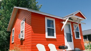 A red wooden house with two windows, a door, and a small porch with two white Adirondack chairs and a grill. - Stylish Studio Cabin in St. Ignace – Near Mackinac Ferry, WiFi & AC (St. Ignace)
