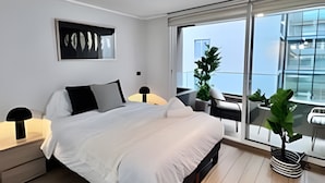 A modern bedroom with a large bed, bedside lamps, a framed artwork, a potted plant, and a balcony with a view. - Fantástico estudio en CLC (Santiago)