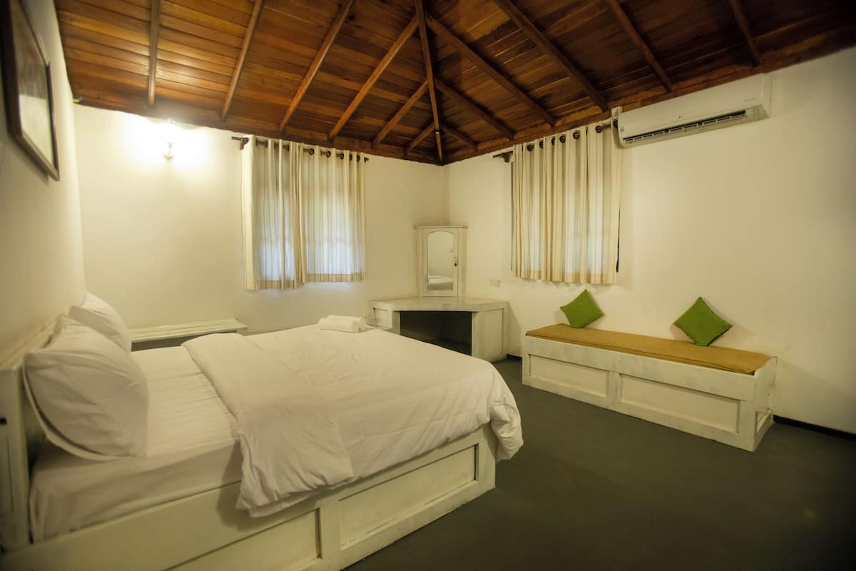 A bedroom with a wooden ceiling, a bed with white bedding, a bench with cushions, a mirror, and an air conditioning unit.