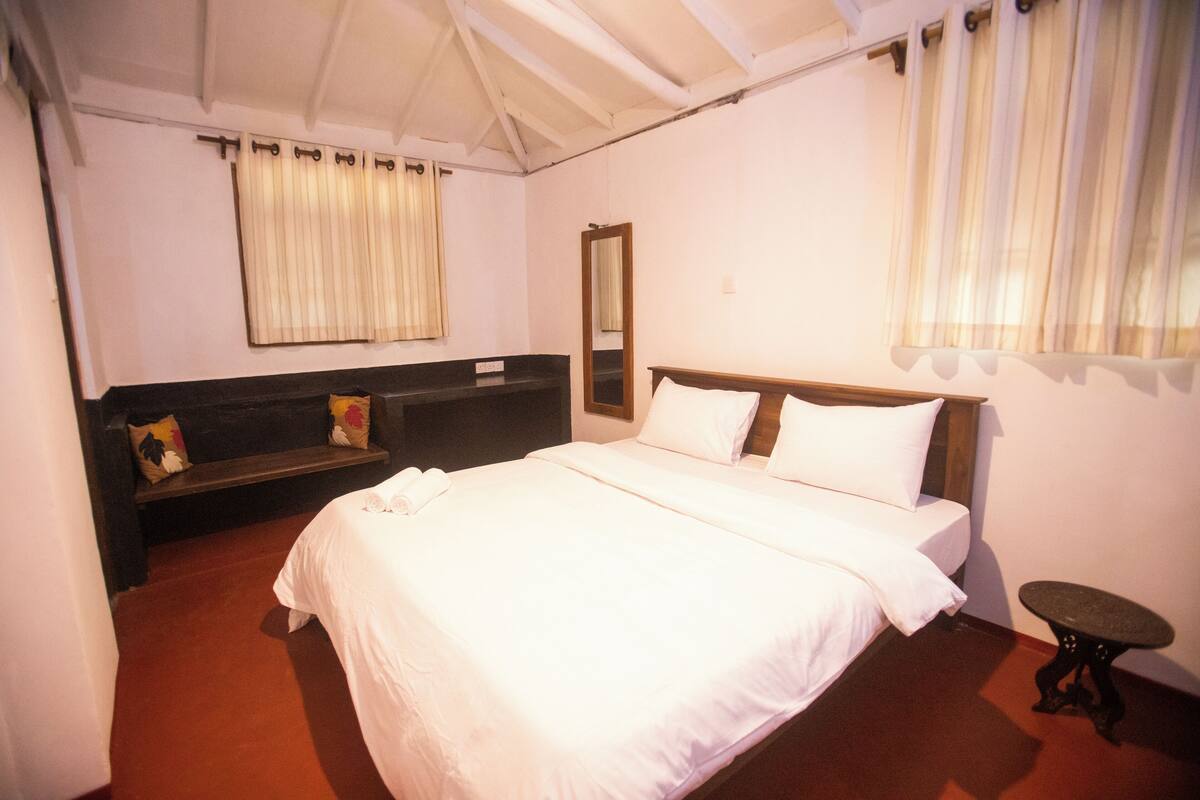 A bedroom with a large bed, a bedside table, a bench with cushions, a mirror, and a window with curtains.