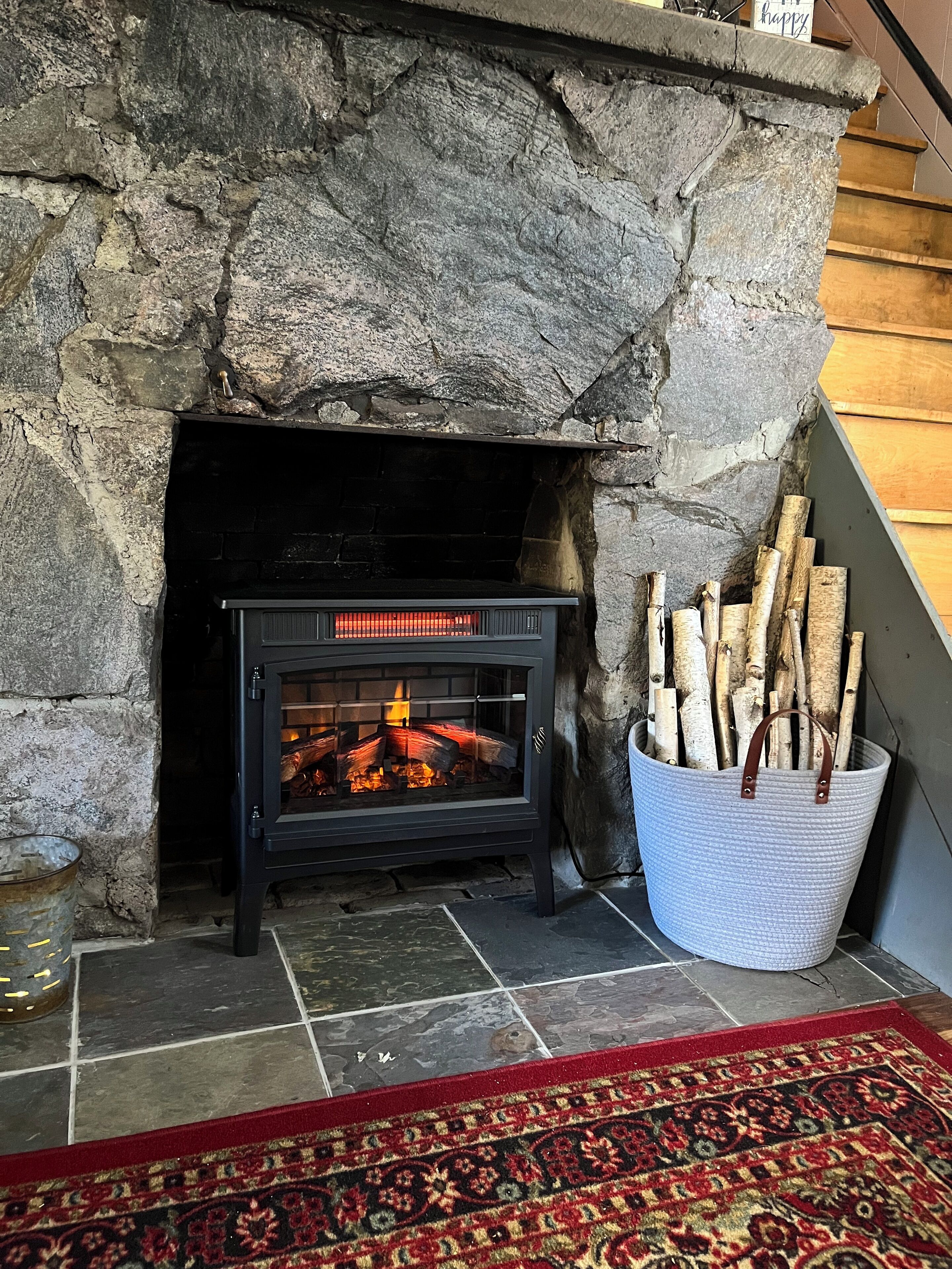 A stone fireplace with a black wood-burning stove, logs in a basket, and a patterned rug on the floor.