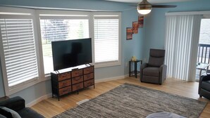 A living room with a flat-screen TV on a wooden cabinet, a grey sofa, a grey armchair, and a ceiling fan. - 2nd Floor Lakeview 2-Bedroom Condo in Geneva U4 (Geneva)