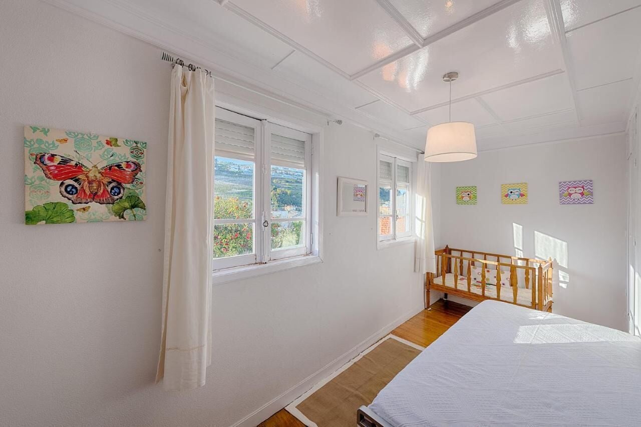 A bedroom with a bed, a wooden crib, a window with curtains, and a butterfly painting on the wall.