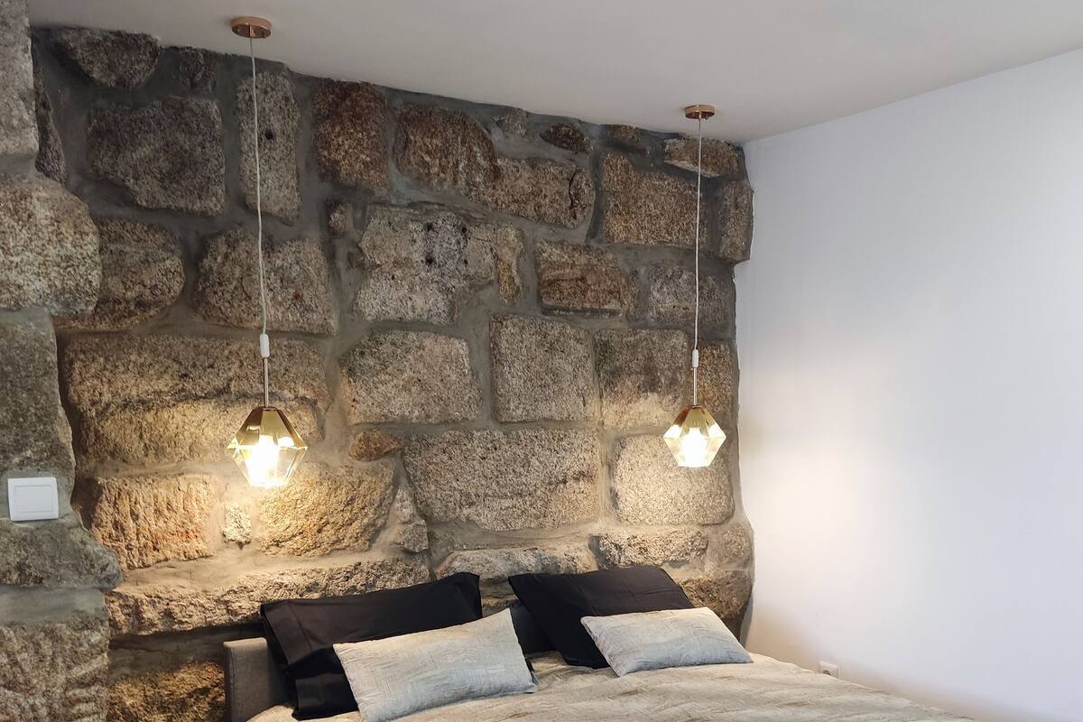Un dormitorio con pared de piedra, cama con manta gris y dos lámparas colgantes.