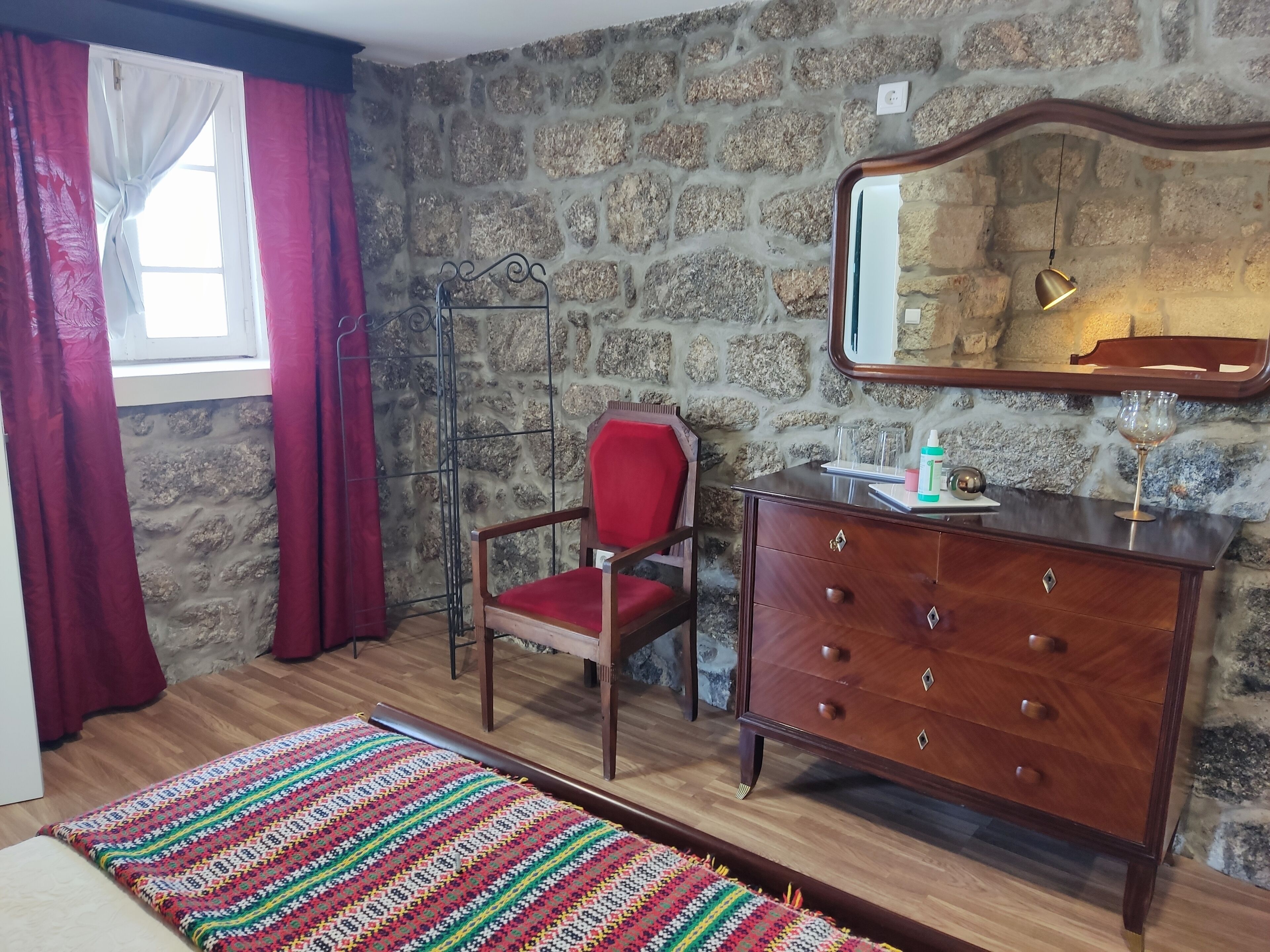 A room with a stone wall, a wooden dresser, a red chair, a patterned rug, and a window with red curtains.