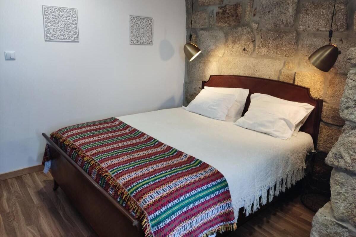Un dormitorio con cama de madera, ropa de cama blanca, una manta colorida y una pared de piedra.