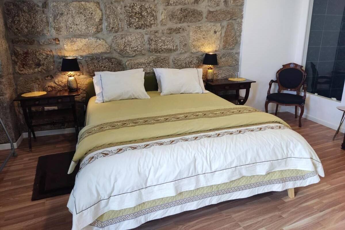 Una cama con colcha blanca y beige, dos mesitas de noche con lámparas, suelo de madera y pared de piedra.