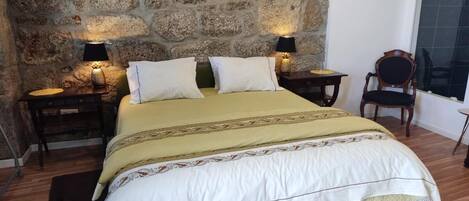 A bed with a white and beige bedspread, two bedside tables with lamps, a wooden floor, and a stone wall.