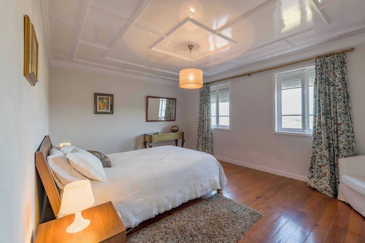 Un dormitorio con cama, lámpara de noche, mesita de madera y una pequeña mesa con lámpara.