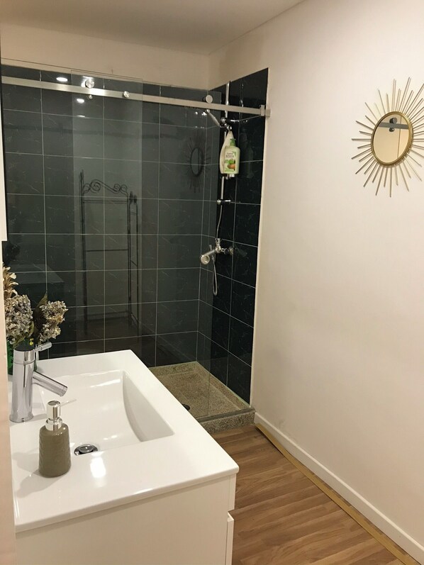 A bathroom with a white sink, a shower with black tiles, and a sunburst mirror on the wall.