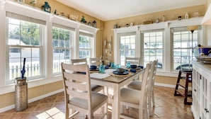 Un comedor con una mesa de madera preparada para cuatro personas, rodeada de sillas. Varias ventanas con marcos blancos y estantes encima de ellas exhiben diversos objetos decorativos.
