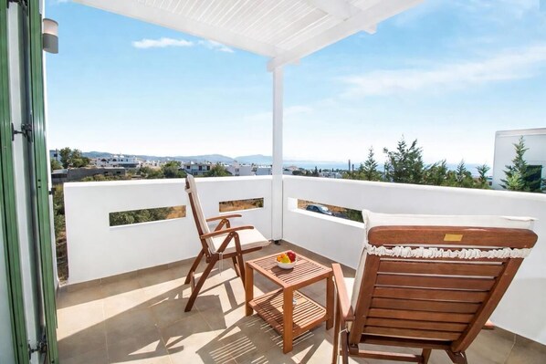 Un balcón con barandilla blanca, dos sillas de madera, una mesita con fruta y vistas de la ciudad.