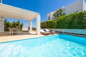 A swimming pool with lounge chairs and a pergola, surrounded by greenery and a white building.