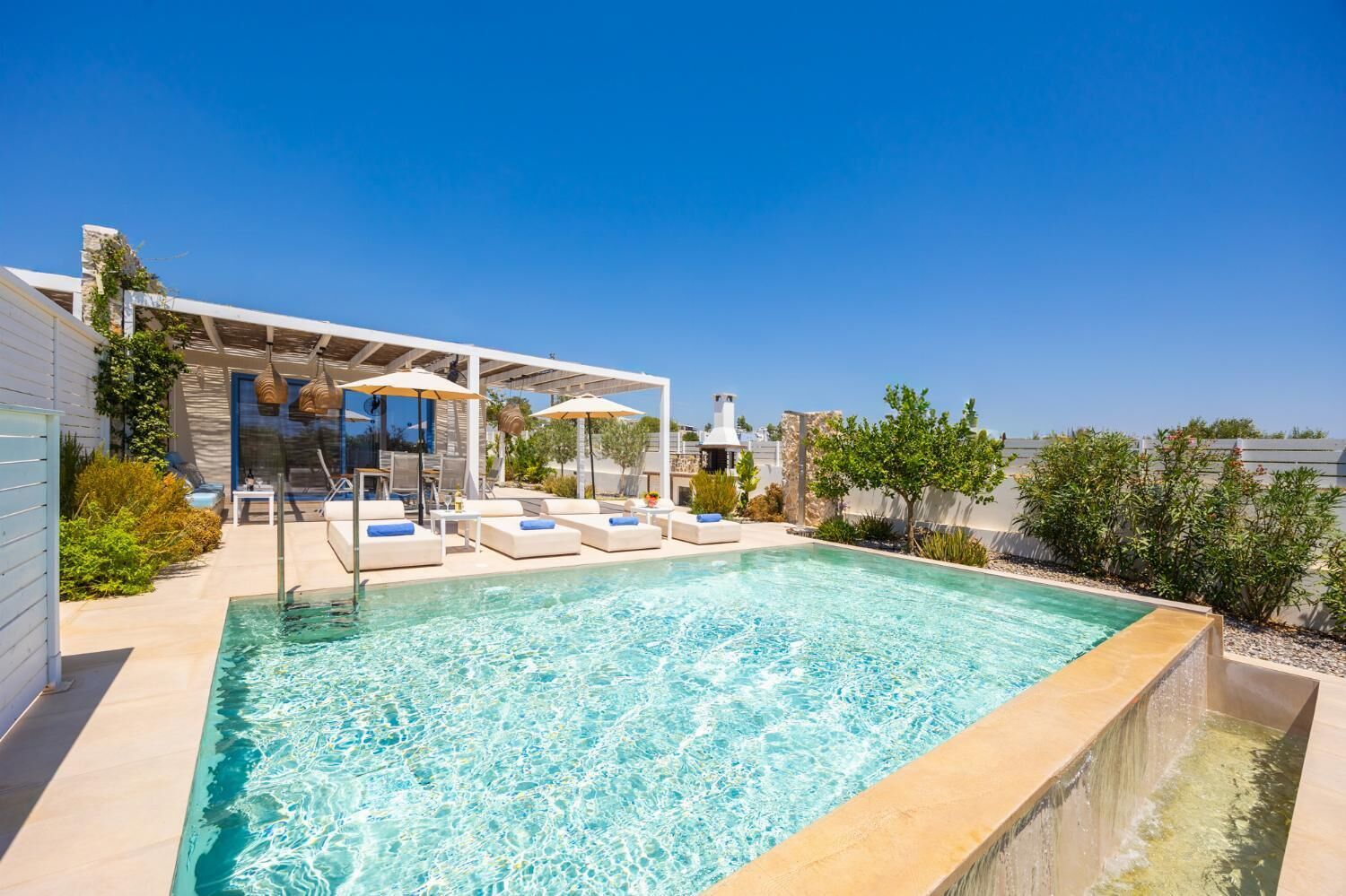 A swimming pool with lounge chairs and umbrellas, surrounded by a patio area with plants and a clear blue sky.