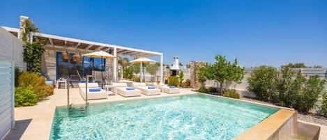 A swimming pool with lounge chairs and umbrellas, surrounded by a patio area with plants and a clear blue sky.