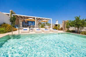 Zona de piscina con sillas de descanso y sombrillas, un muro de piedra y un cielo azul despejado.