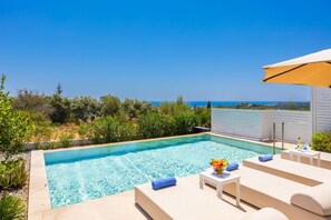 A rectangular pool with clear blue water, surrounded by lounge chairs and a small table with a bottle and fruit.