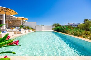 Una piscina con sillas de descanso y sombrillas, rodeada de vegetación y un muro de piedra.