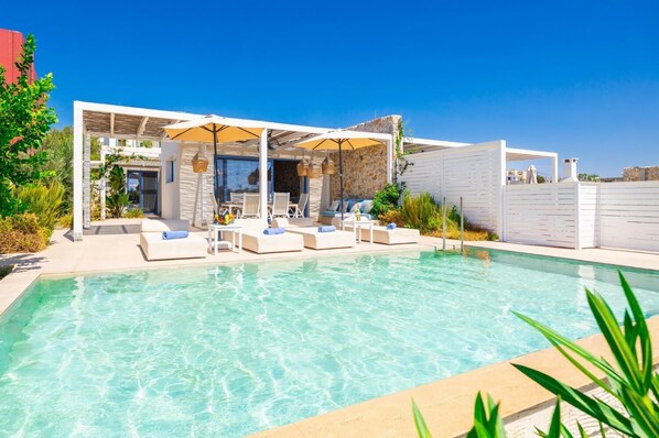 A swimming pool with lounge chairs and umbrellas, surrounded by a white fence and vegetation.