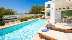 Une piscine entourée d’une clôture blanche et de verdure, avec des chaises longues et une table à proximité.