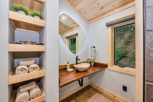 Un baño con estantes de madera, un espejo redondo, encimera de madera, un lavabo y una ventana con vistas a la vegetación.