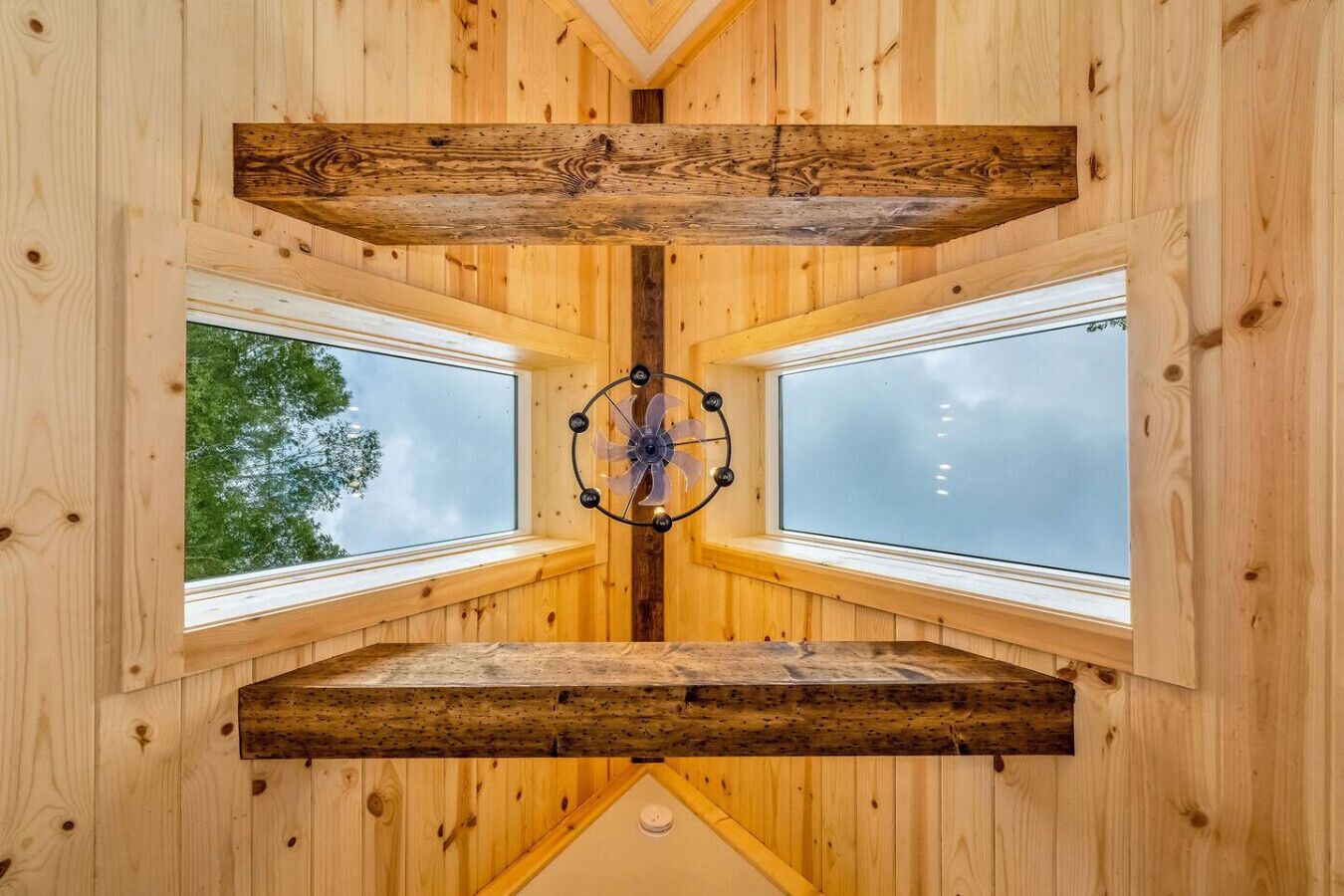 A room with wooden walls and a central ceiling fan. There are four windows on the walls, each with a view of trees.