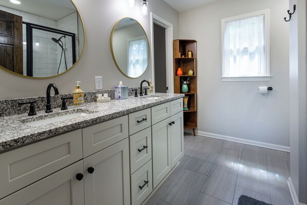 Une salle de bain moderne avec un comptoir en granit, un lavabo double, un grand miroir et une étagère en bois agrémentée d’objets décoratifs.