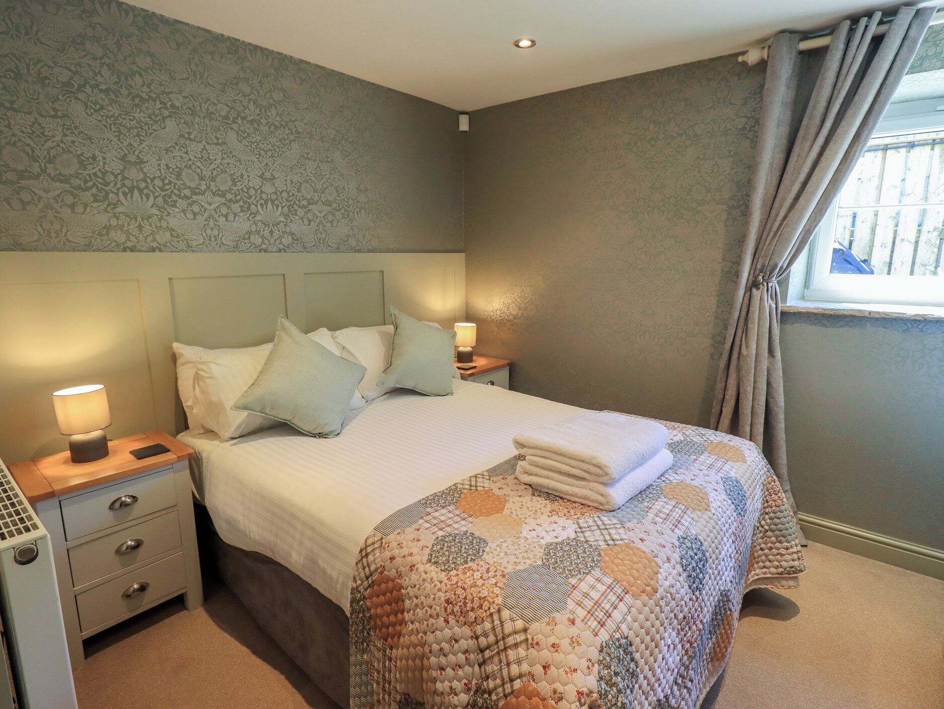 A neatly made bed with pillows, a bedside lamp, a nightstand with drawers, and a window with curtains.