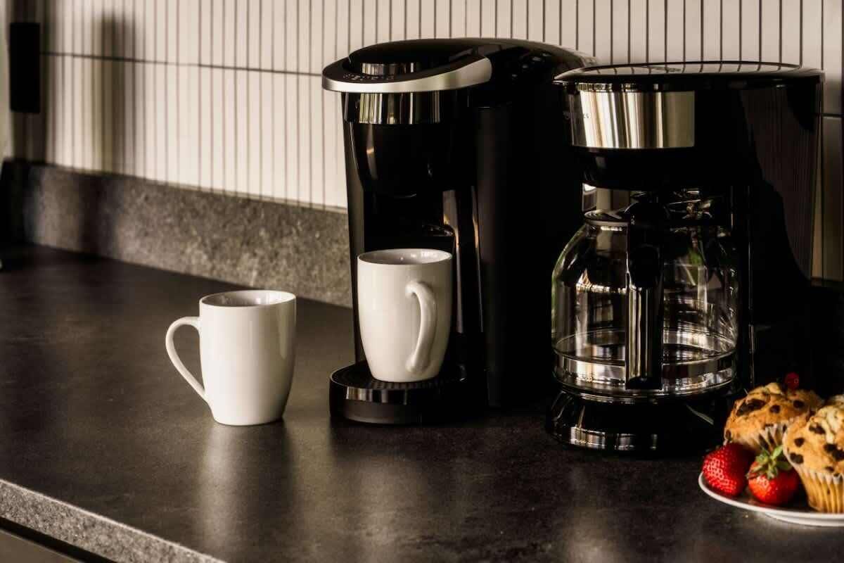 A kitchen counter with a coffee maker, two white mugs, and a plate of muffins.