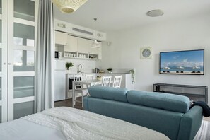 A modern bedroom with a bed, a teal sofa, a dining area with chairs, and a kitchen in the background.