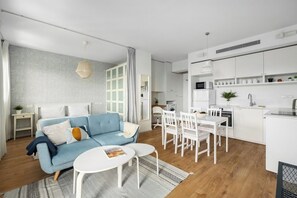 A modern living room with a blue sofa, white dining table, and chairs, a kitchen with white cabinets, and a grey patterned wall.
