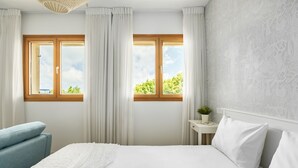 Ein Schlafzimmer mit einem großen Bett, zwei Fenstern mit Vorhängen, einem Nachttisch mit einer Pflanze und einem blauen Sessel.