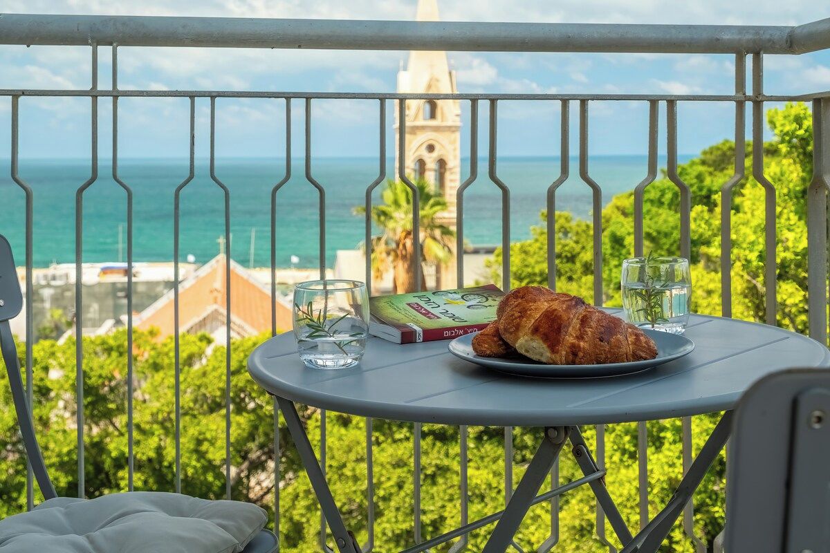 A balcony with a table set for a meal, overlooking a coastal view with a church spire in the distance.