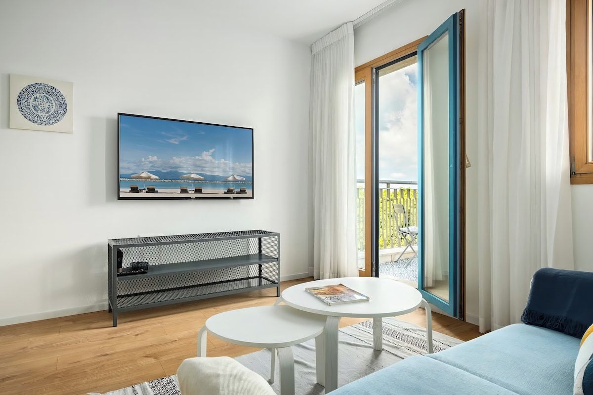 A modern living room with a large TV, a sofa, a coffee table, and a balcony with a view of the sea.
